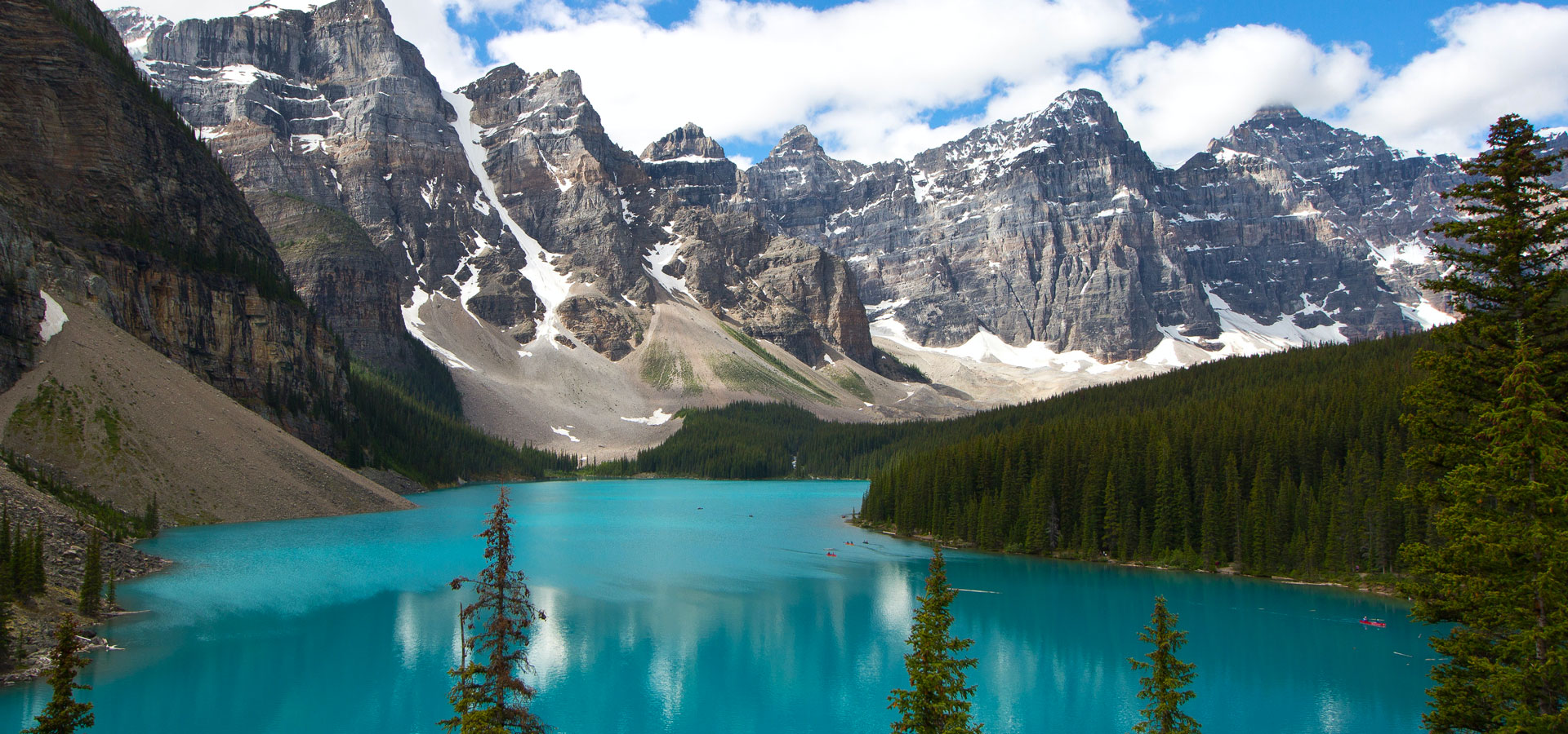 A glacial fed lake of vivid turquoise with lone standing pines in the foreground. In the distance are ice capped mountains enveloping the lake and a blue sky with snow white clouds. 