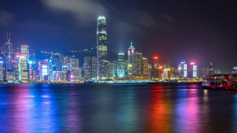 A view across Victoria Harbour toward the city of Hong Kong at night. The bright lights of the Skyscrapers illuminate several different colours across the water.