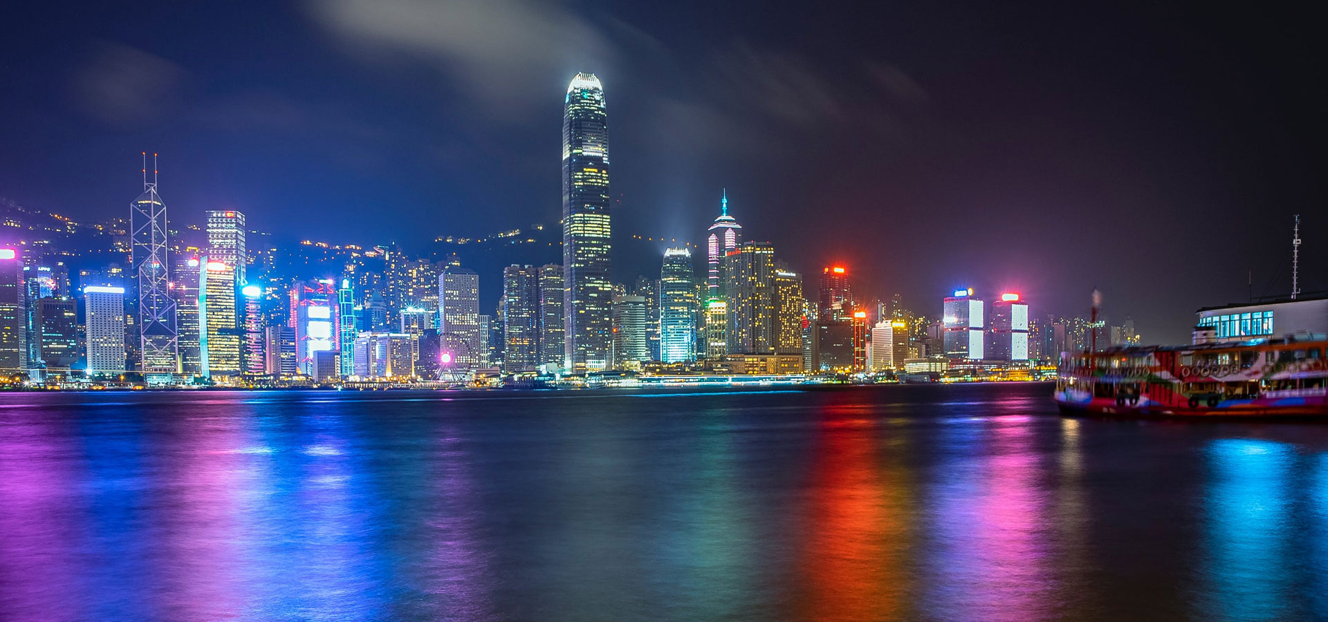 A view across Victoria Harbour toward the city of Hong Kong at night. The bright lights of the Skyscrapers illuminate several different colours across the water.