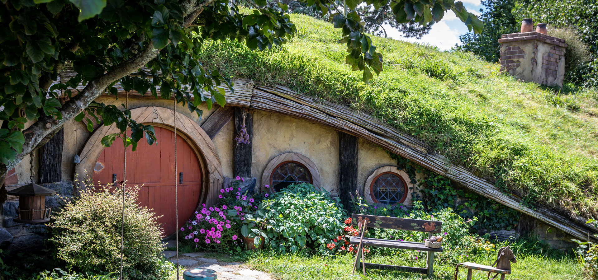 A Hobbit House set into the side of a grassy topped hill with a large round brown door and circular windows. The front garden is highlighted by green shrubs, colourful flowers and a wooden bench seat in the sunshine.