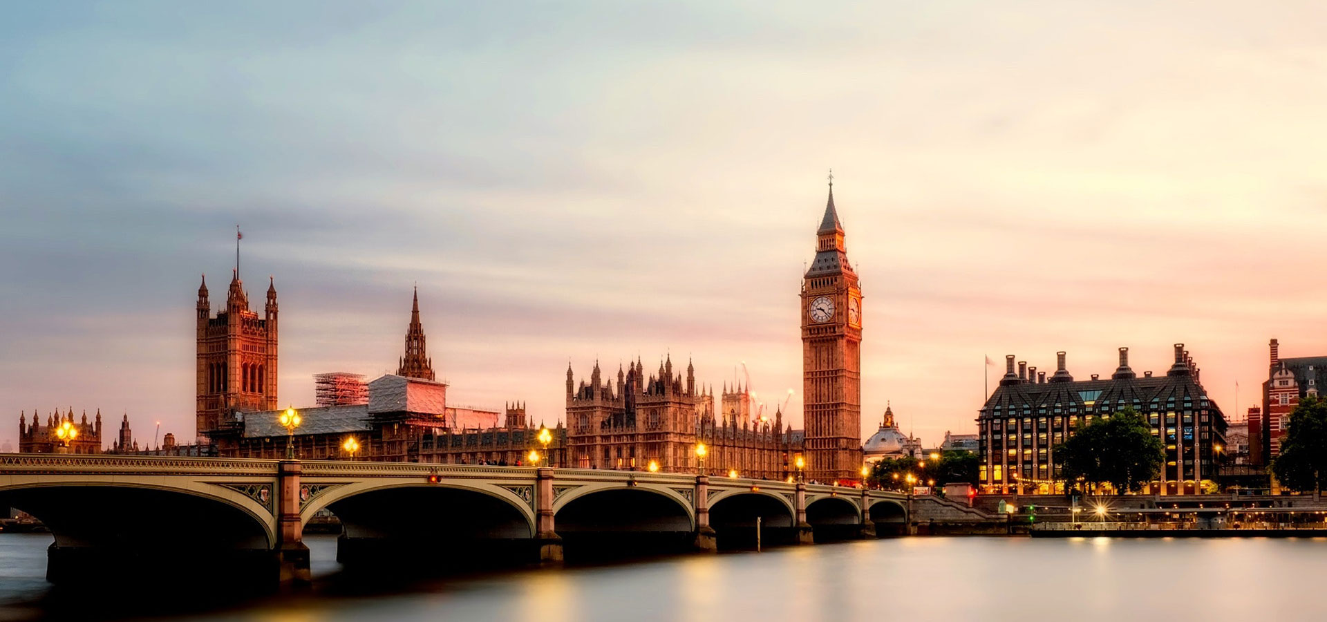 View across the Thames River overlooking West Minster Bridge and Big Ben, against pink skies at dusk.