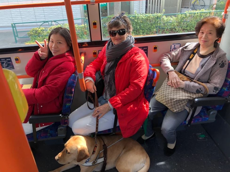 Nixon laying down by Nina's feet on the bus with Nina and nearby passengers smiling