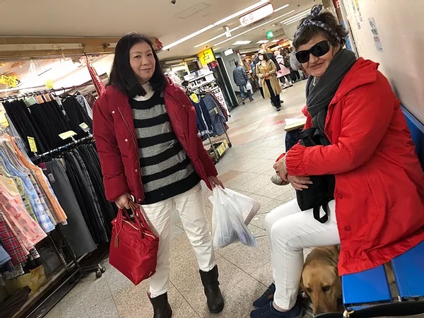 Nina and her friend outside a shop in Japan with Guide Dog Nixon tucked under her feet out of the way