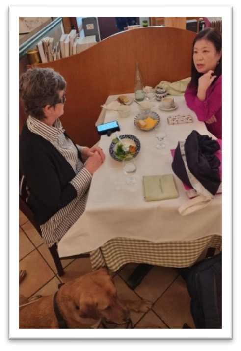 Nina sits at a restaurant table with a member of her Japanese host family. At her feet lies Assistance Dog Nixon wearing his identifying harness.