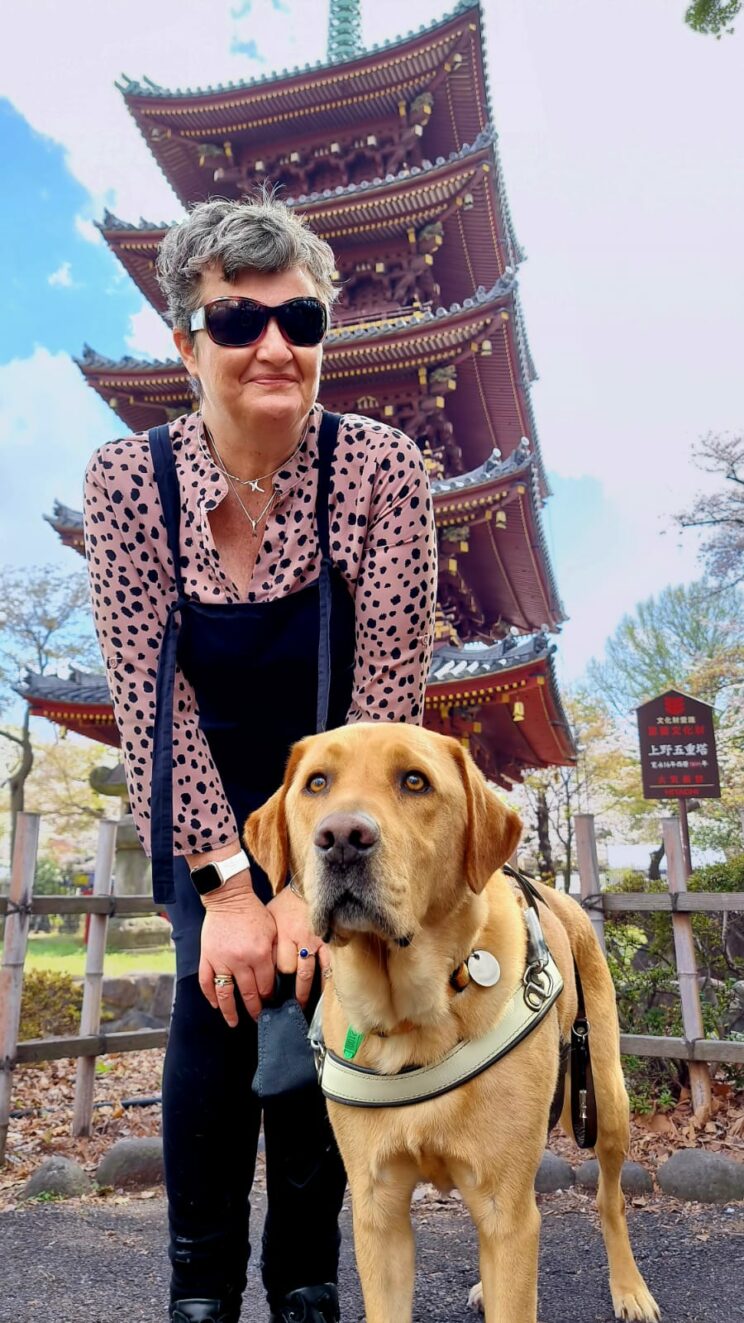 Nina Smith, TravelPaws Founder & Director, stands in front of a 5-story pagoda in Tokyo wearing a black pinafore over a pink and black spotty top. Still looking at the camera, she leans down slightly to be closer to Assistance Dog Nixon, a golden Labrador, who stands beside her wearing his identifying harness.