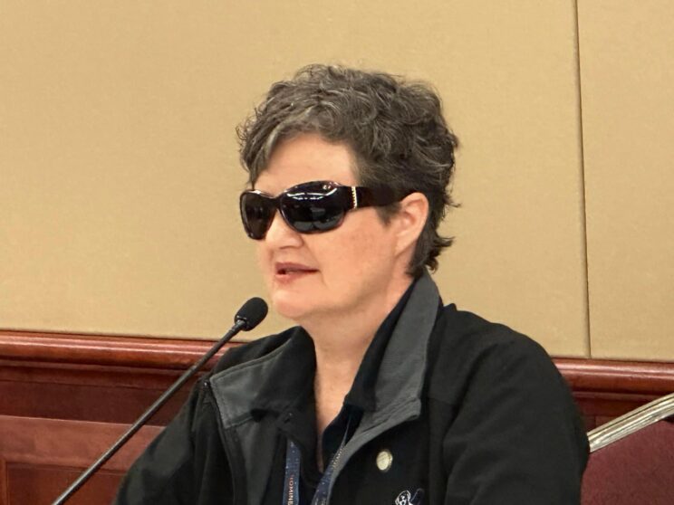 AdvocacyWebpageImage_TaxiRefusalsPresentation Close up image of TravelPaws Founder & Director, Nina Smith, talking into a microphone. Nina wears a black jacket with the TravelPaws logo, which is a graphic of an Assistance Dog in harness next to a suitcase.