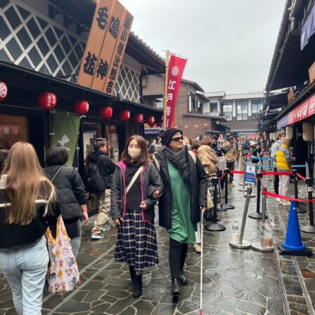 Nina walks with her arm looped through Mitaru's elbow. She holds her cane out, scanning the ground in front of her. The pair stroll through a bustling shopping street, having just passed a roped-off section.