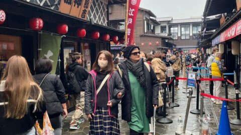 Nina walks with her arm looped through Mitaru's elbow. She holds her cane out, scanning the ground in front of her. The pair stroll through a bustling shopping street, having just passed a roped-off section.