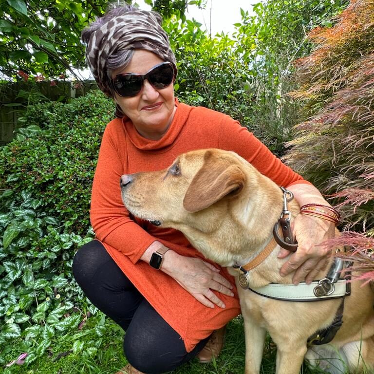 TravelPaws Founder & Director, Nina Smith, crouches next to Guide Dog Nixon, a golden Labrador. Nina wears a long sleeve orange dress over black leggings with a leopard print bandana in her hair. Nixon wears his brown and white identifying harness. The pair are surrounded by greenery.